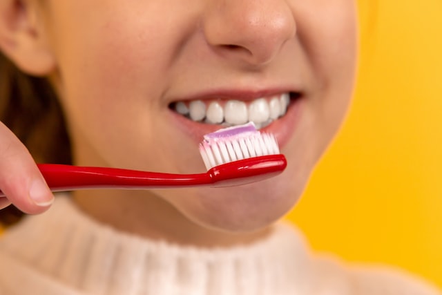 photo of lower half of person's face, white teeth visible. toothbrush with toothpaste held in front of mouth. yellow background